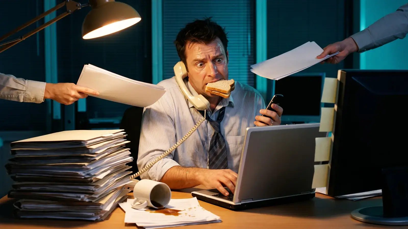 Profissional sobrecarregado à mesa do escritório, à noite: fala ao telefone com o fone preso no ombro, morde um sanduíche, digita no notebook e segura um celular, enquanto duas mãos lhe estendem mais papéis por cima de pilhas de pastas